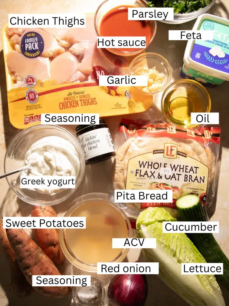 Ingredients for buffalo chicken pitas including chicken thighs, hot sauce, Greek yogurt, feta cheese, pita bread, sweet potatoes, cucumber, red onion, garlic, olive oil, and seasoning.