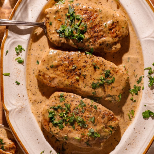 Peppercorn crusted pork chops covered in creamy parmesan sauce and finished with fresh parsley on a serving platter.