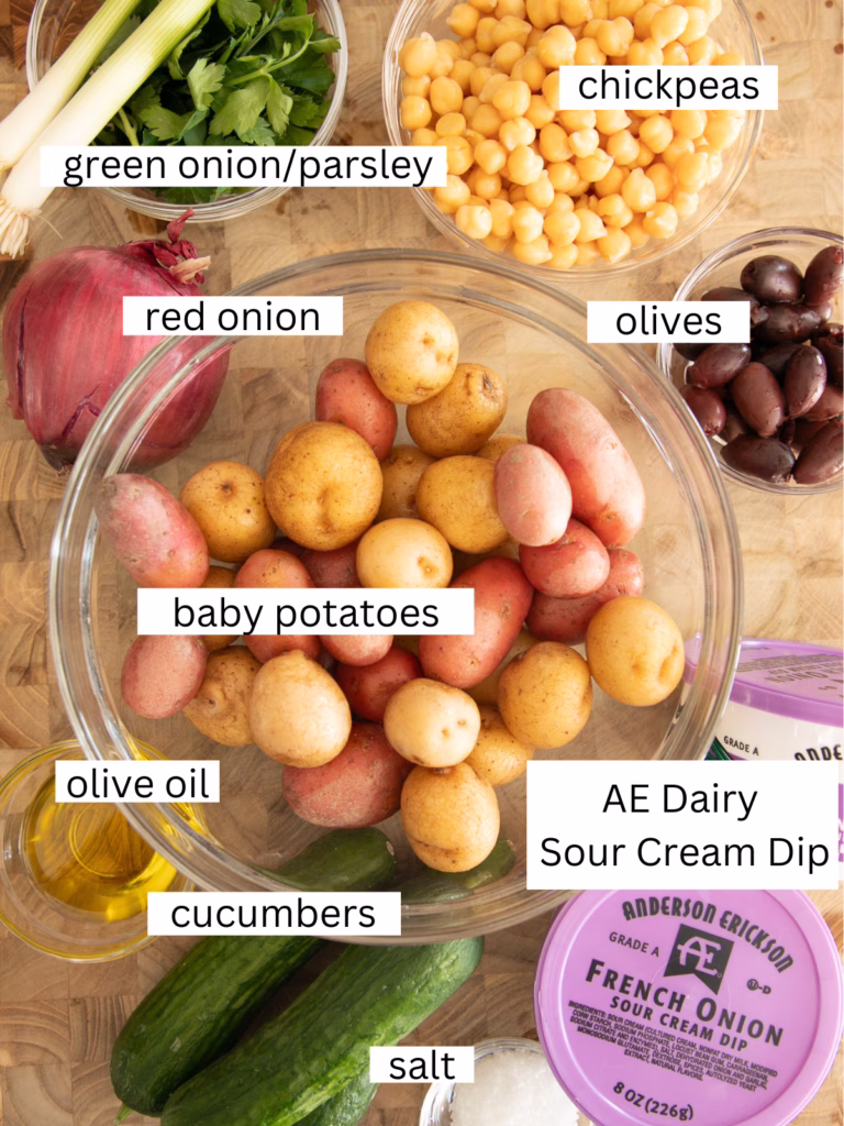 Overhead view of baby potatoes, chickpeas, cucumbers, red onion, olives, parsley, green onion, olive oil, salt, and sour cream dip arranged on a wooden surface.