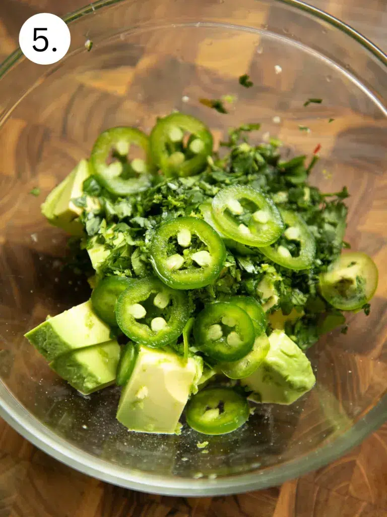 Avocado chunks with sliced jalapeños, fresh cilantro, and lime juice in a glass bowl before blending into sauce.