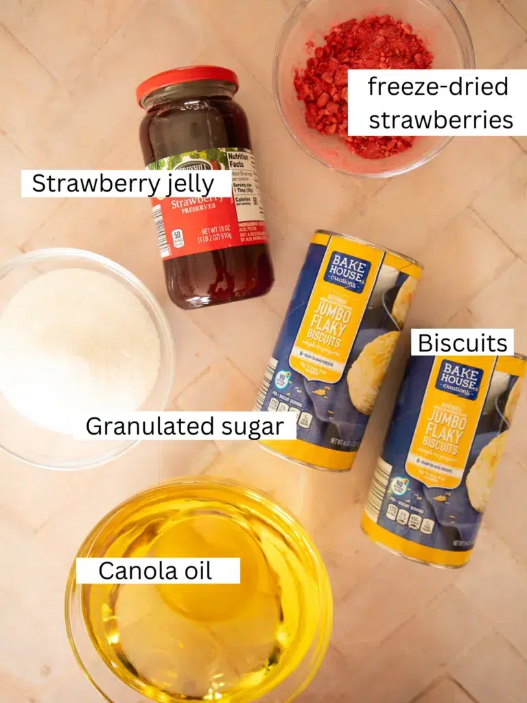 Ingredients for Valentine’s Day biscuit donuts including canned biscuits, strawberry jelly, freeze-dried strawberries, granulated sugar, and canola oil arranged on a countertop.