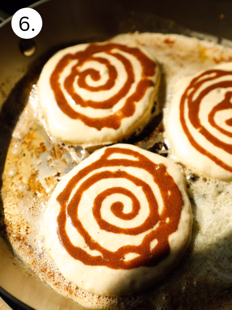 Buttermilk pancakes cooking in a skillet with a cinnamon swirl pattern forming on top