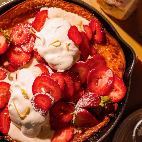 Close-up of a strawberry honey butter Dutch baby pancake with sliced strawberries, whipped cream, and a vintage serving spoon.