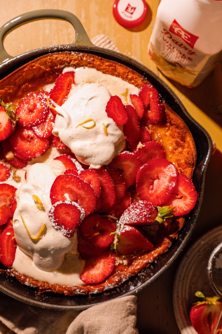 Close-up of a strawberry honey butter Dutch baby pancake with sliced strawberries, whipped cream, and a vintage serving spoon.