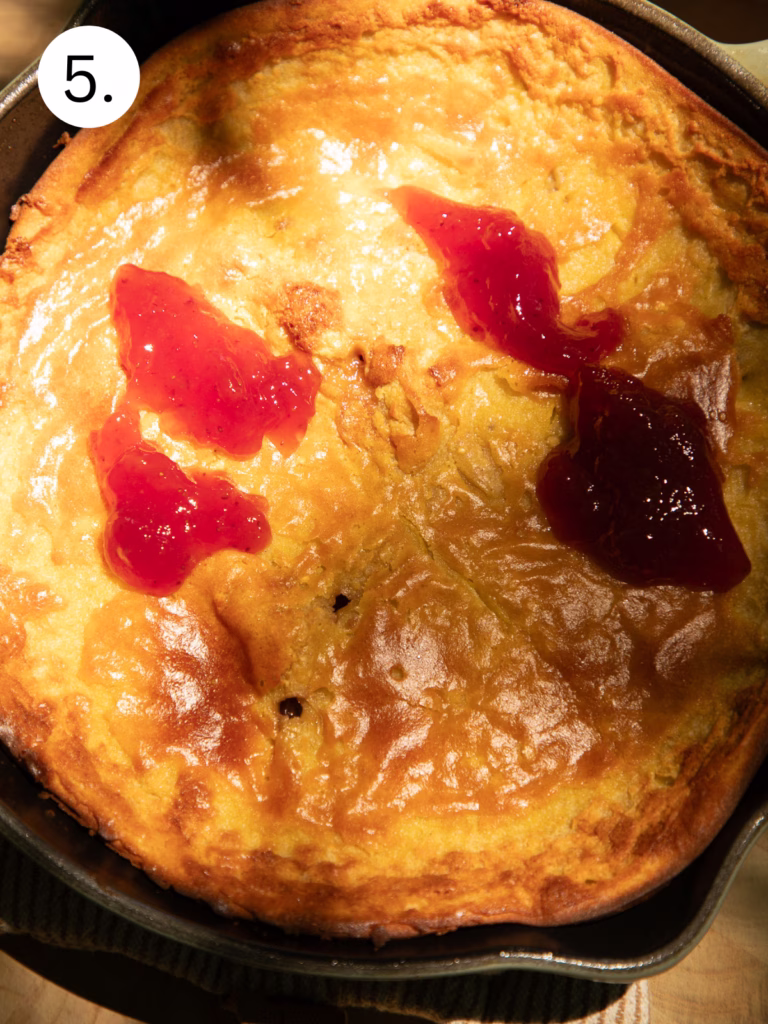 Golden baked Dutch baby pancake in a cast iron skillet topped with spoonfuls of strawberry jelly.