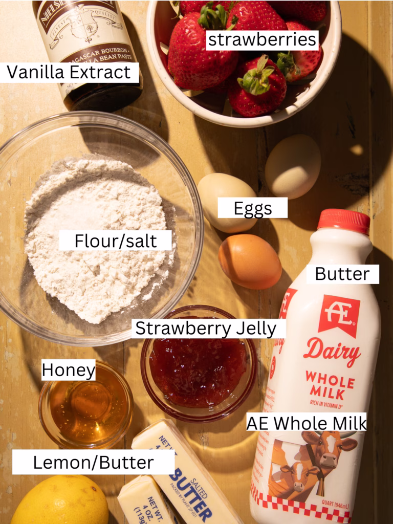 Ingredients for a strawberry honey butter Dutch baby skillet pancake including strawberries, eggs, flour, vanilla, honey, butter, strawberry jelly, lemon, and AE Dairy whole milk arranged on a wooden table.