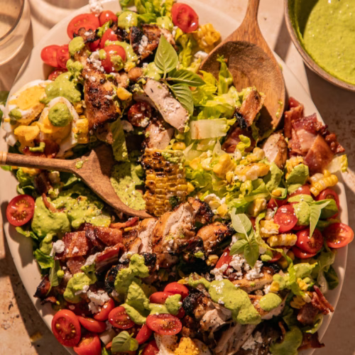 Close-up of a Grilled Green Goddess Cobb Salad served with grilled chicken, charred corn, bacon, feta, tomatoes, eggs, fresh basil, and creamy Green Goddess dressing.