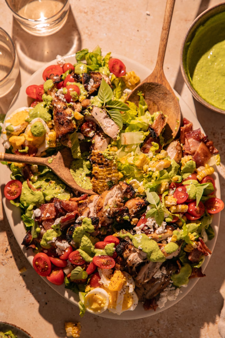 Close-up of a Grilled Green Goddess Cobb Salad served with grilled chicken, charred corn, bacon, feta, tomatoes, eggs, fresh basil, and creamy Green Goddess dressing.