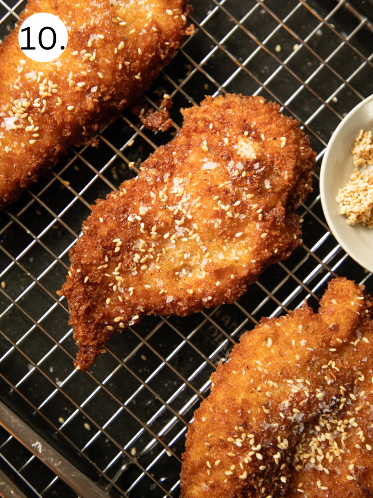 Golden brown crispy chicken cutlets topped with sesame seeds and flaky salt resting on a wire rack after frying.
