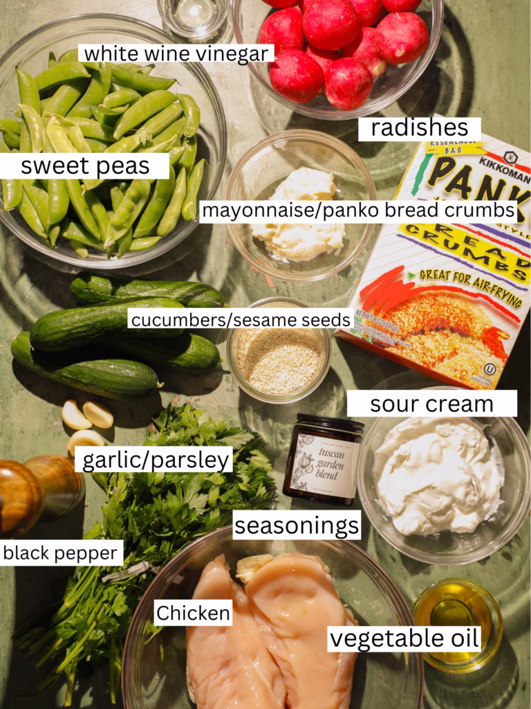 Ingredients for crispy sesame chicken cutlets and creamy sweet pea salad arranged on a table, including chicken breasts, snap peas, cucumbers, radishes, panko breadcrumbs, mayonnaise, sour cream, parsley, garlic, sesame seeds, white wine vinegar, oil, and seasonings.